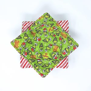 May include: A square, quilted bowl cozy with a green Grinch print, featuring the phrases "Feelin' Grinchy" and "Naughty Nice". The cozy sits atop a red and white striped square. Christmas-themed kitchenware.
