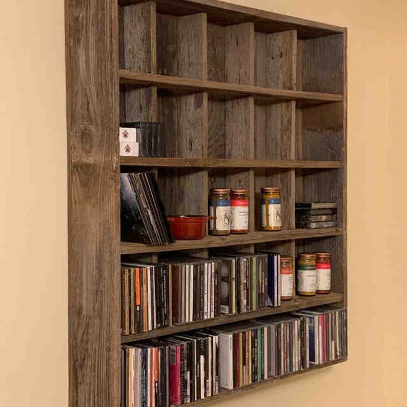 Puede incluir: Estanter&iacute;a de pared r&uacute;stica de madera con m&uacute;ltiples compartimentos. Los estantes est&aacute;n llenos de CDs, discos de vinilo y tarros. La madera tiene un acabado envejecido gris-marr&oacute;n, d&aacute;ndole una est&eacute;tica vintage.