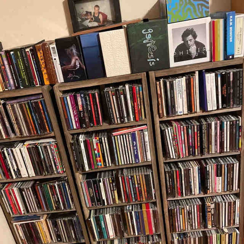Puede incluir: Estanter&iacute;as de madera llenas de una gran colecci&oacute;n de CDs. Los lomos de los CDs son visibles, mostrando una variedad de colores y t&iacute;tulos. Algunas portadas de &aacute;lbumes son visibles, incluyendo una con la palabra "Genesis". Una foto enmarcada se encuentra encima.