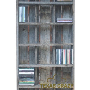 Puede incluir: Estanter&iacute;a r&uacute;stica de madera para CD con m&uacute;ltiples estantes, que muestra varios CD. La estanter&iacute;a est&aacute; hecha de madera envejecida con una mezcla de tonos grises y marrones. El texto "TEXAS CRAZY" es visible en la parte delantera.