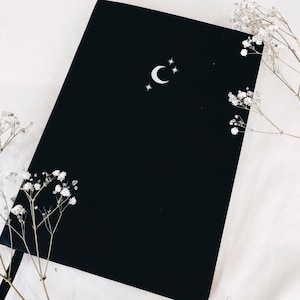 Könnte beinhalten: Ein schwarzes Notizbuch mit einem Halbmond- und Sternenmuster. Das Notizbuch liegt auf einer weißen Oberfläche mit weißen Blumen.