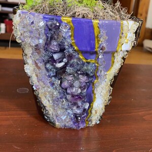 Puede incluir: Una maceta de color morado y dorado con un diseño de geoda de cristal. La maceta está llena de musgo y una pequeña planta verde.