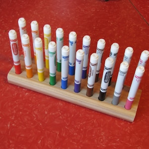 May include: A wooden marker holder with a variety of Crayola markers in different colors. The markers are arranged in a neat row, with the colors visible. The holder is made of light-colored wood and sits on a red surface.
