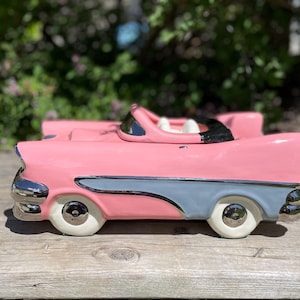 May include: A pink ceramic vintage car planter with chrome accents and white tires. The car features a light blue stripe along the side and a black windshield. The planter is designed to resemble a classic convertible.