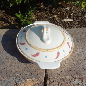 Pu&ograve; includere: Piatto da forno in ceramica bianca con coperchio. Il coperchio ha un manico e un design decorativo di peperoncini rossi, triangoli blu e linee gialle su uno sfondo crema.