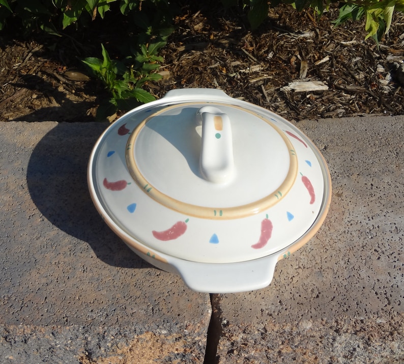 Pu&ograve; includere: Piatto da forno in ceramica bianca con coperchio. Il coperchio ha un manico e un design decorativo di peperoncini rossi, triangoli blu e linee gialle su uno sfondo crema.