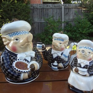 May include: Three ceramic pig figurines dressed as chefs. The pigs wear white chef hats with blue checkered bands, bow ties, and checkered jackets. One holds a plate, another a teapot, and the third a teacup. The figurines are on a brown surface.