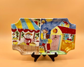 May include: A colorful ceramic serving tray with a whimsical scene of people shopping at a market. The tray features a yellow awning, a red building, and a blue sky with white clouds. The text "CAMMISA" is visible on the tray.