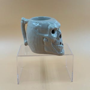 May include: A gray ceramic mug shaped like a human skull. The mug features a handle designed to resemble a bone. The skull has detailed facial features, including eye sockets and a nose cavity. The mug is displayed on a clear acrylic stand.