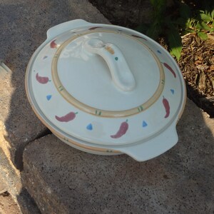 Pu&ograve; includere: Piatto da forno in ceramica bianca con coperchio. Il coperchio ha una maniglia e un design di peperoncini rossi, triangoli blu e foglie verdi su uno sfondo beige.