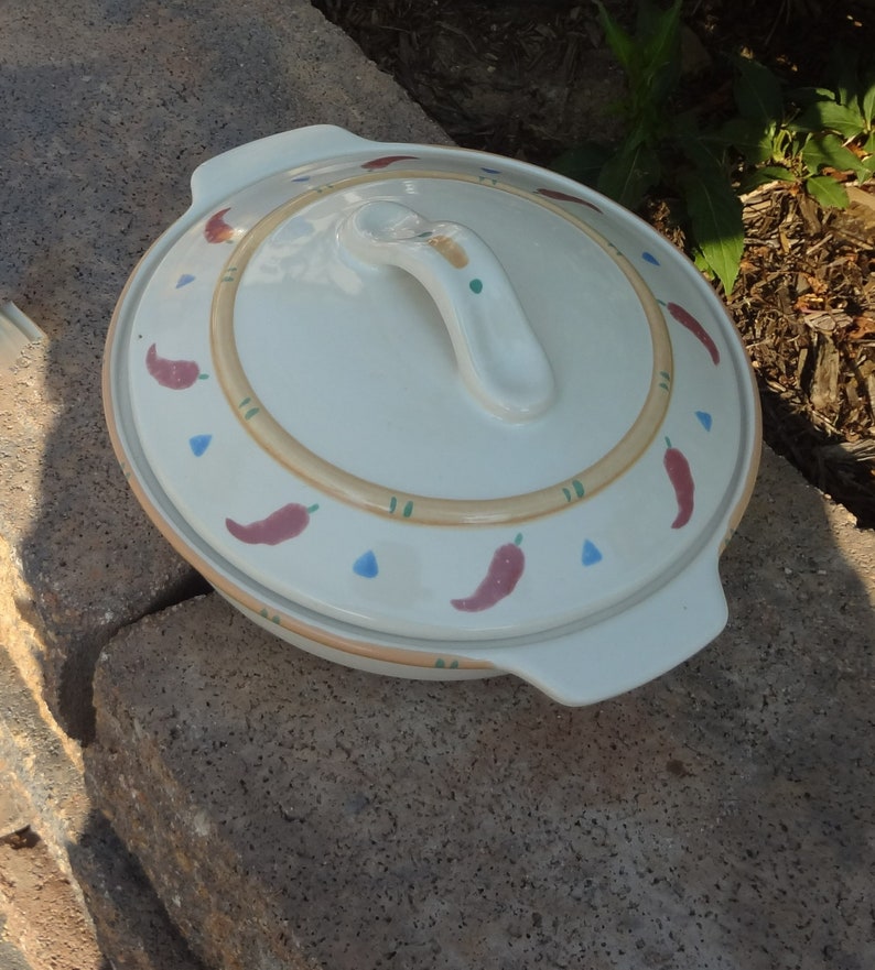Pu&ograve; includere: Piatto da forno in ceramica bianca con coperchio. Il coperchio ha una maniglia e un design di peperoncini rossi, triangoli blu e foglie verdi su uno sfondo beige.