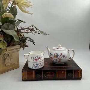 May include: A white porcelain tea set with a teapot and creamer. The teapot and creamer are decorated with colorful floral and bird designs and gold trim. The set is displayed on a vintage book.