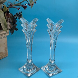 May include: Two clear glass candle holders with a decorative, layered design. The candle holders have a square base and a tall, slender stem that flares out at the top. The background is a solid blue.