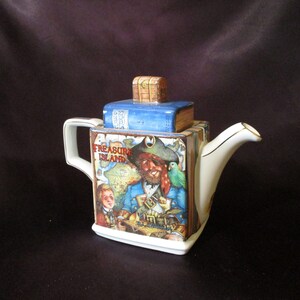 May include: A white ceramic teapot shaped like a book with a blue and white cover. The book is titled "Treasure Island" and features a pirate with a parrot on his shoulder. The teapot has a gold handle and spout.