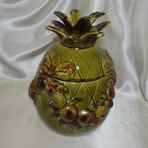 May include: A green ceramic pineapple cookie jar with brown accents and a detailed pineapple design. The jar is decorated with brown fruit and leaves around the base.