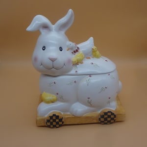 May include: White ceramic bunny cookie jar with yellow chicks and a floral design. The bunny is on wheels with black and white checkered wheels.