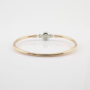 May include: A gold-coloured bangle bracelet with a silver-coloured clasp. The bracelet has a simple, elegant design, with a smooth, rounded band. The clasp is circular and appears to be made of metal. The bracelet is displayed on a white surface.