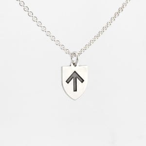 May include: A silver necklace featuring a shield-shaped pendant with a black arrow pointing upwards. The pendant hangs from a delicate chain. The jewelry is set against a plain background.