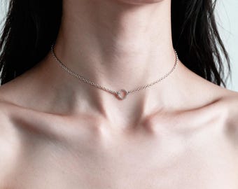 May include: A silver-toned choker necklace featuring a delicate chain with small, uniform circular links. The necklace is centered by a single, small, circular ring pendant that connects the two sides of the chain, resting against the base of the neck.
