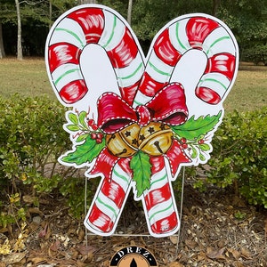 Puede incluir: Un cartel de jard&iacute;n navide&ntilde;o de bast&oacute;n de caramelo rojo y blanco con un lazo rojo y campanas doradas. El cartel est&aacute; hecho de madera y tiene una rama de acebo verde con bayas rojas. El cartel est&aacute; decorado con un patr&oacute;n de rayas blancas y verdes. El cartel est&aacute; hecho por DREZ Artwork & Design.