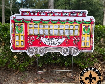 Könnte beinhalten: Ein rotes und gelbes Weihnachtsgartenschild mit einem Straßenbahn-Motiv und dem Schriftzug „Merry Christmas“ auf einem Banner. Die Straßenbahn ist mit Lichtern, Girlanden und Zuckerstangen geschmückt. Das Schild steht auf einem Metallpfahl.