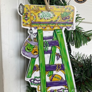 May include: A Mardi Gras themed Christmas ornament featuring a green and purple ladder with a yellow mailbox on top. The mailbox has the words "Our Krewe" on it. The ornament is decorated with beads, masks, and other Mardi Gras symbols.