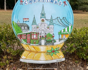 Könnte beinhalten: Ein handbemalter Weihnachts-Schneekugel aus Holz mit blauem Hintergrund und einer Szene der St. Louis Cathedral in New Orleans. Die Schneekugel ist mit roten Beeren und einem grünen Stechpalmenzweig verziert. Der Text "Merry Christmas" ist in roten und weißen Buchstaben auf der Oberseite der Schneekugel geschrieben.