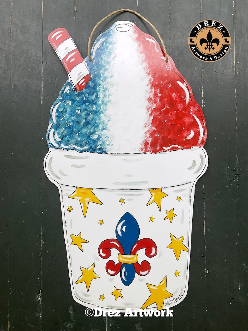 Op de afbeelding: Een rode, witte en blauwe sneeuwkegel met een rood-wit gestreepte rietje in een witte beker met gouden sterren en een blauwe fleur-de-lis. De beker heeft de tekst "Drez Artwork" op de bodem.