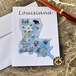May include: A white card with a blue outline of the state of Louisiana with various illustrations of things associated with the state, including a pelican, a magnolia flower, a strawberry, a crawfish, and oysters. The text "Louisiana" is at the top of the card. The card also has a small gold circle with the text "DREZ Artwork & Design" in black.