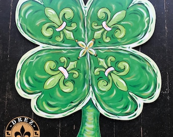 May include: A green four-leaf clover with white and gold fleur-de-lis designs. The clover is painted on a wooden surface and has a brown rope hanger. The text "@DREZ ARTWORK AND DESIGN" is visible at the bottom of the image.
