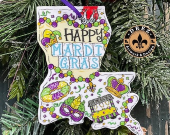 Puede incluir: Un adorno blanco en forma de Luisiana con las palabras "Happy Mardi Gras" en negro y azul. El adorno presenta símbolos coloridos del Mardi Gras como una máscara, una corona y cuentas. Se adjunta una cinta morada para colgar.