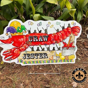 May include: A hand-painted Mardi Gras crawfish sign with the words "Craw Jester" and a jester hat on the crawfish. The sign is surrounded by silhouettes of people and beads.