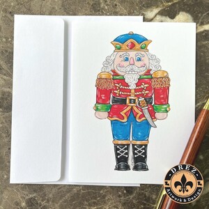 May include: A hand-drawn greeting card featuring a colorful nutcracker soldier. The nutcracker wears a red jacket, blue pants, and a blue and gold crown. A white envelope is to the left of the card. A pen and a sticker are in the bottom right corner.