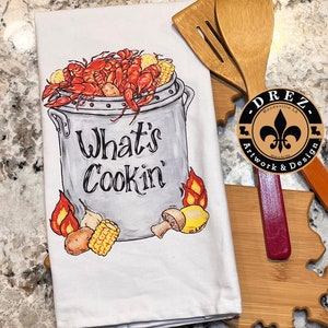 May include: White kitchen towel with a colorful illustration of a pot of crawfish with the text "What's Cookin'" and a logo for Drez Artwork & Design.