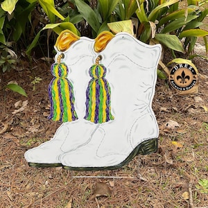 May include: White boots with gold and green tassels. The boots are painted with a Mardi Gras theme. The boots are on a wooden stand. The text "DREZ Artwork & Design" is on a gold circle.