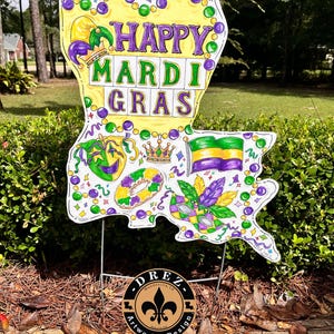 May include: A yellow and purple Mardi Gras sign shaped like the state of Louisiana. The sign features the text "Happy Mardi Gras" and various Mardi Gras symbols, including masks, beads, and a king cake. The sign is on a metal stake and is ready to be placed in the ground.