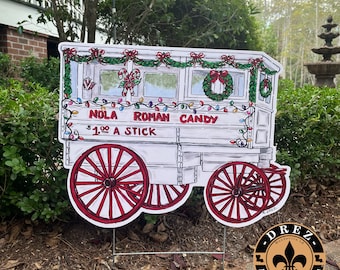 Könnte beinhalten: Ein weißer Holzwagen, der mit roter und grüner Girlande, Zuckerstangen und Lichtern für Weihnachten dekoriert ist. Der Wagen hat ein Schild mit der Aufschrift "NOLA ROMAN CANDY 1,00 € PRO STANGE".