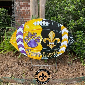 May include: A painted wooden football with a purple and yellow tiger paw print on one side and a black fleur-de-lis on the other side. The words "Tigers" and "Saints" are written on the football. The football is on a wooden stake.