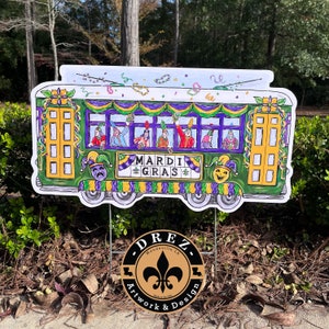 May include: A Mardi Gras-themed yard sign featuring a green trolley car with gold doors and purple accents. The sign displays the words "Mardi Gras" and includes illustrations of people celebrating. A round sign with the text "DREZ Artwork & Design" is at the bottom.