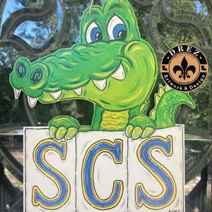 May include: A green alligator-shaped wooden sign with the letters "SCS" in blue and yellow. The alligator has blue eyes and white teeth. The sign is hanging by a string and has a logo that says "DREZ Artwork & Design".