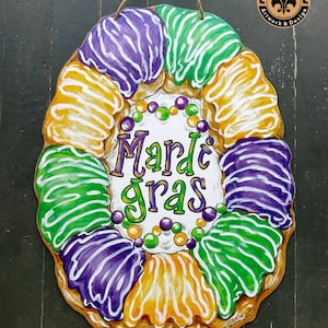 May include: A Mardi Gras wreath made of a painted wooden donut with purple, green, and gold icing. The wreath has a white sign in the center that says "Mardi Gras".