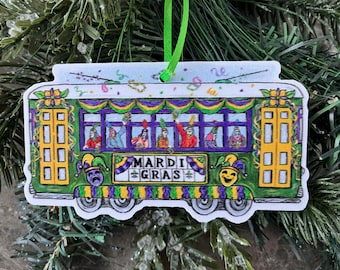 Puede incluir: Un adorno de tranvía de Mardi Gras verde y amarillo con un ribete de Mardi Gras morado, dorado y verde. El tranvía tiene un letrero que dice "Mardi Gras" y tiene gente en las ventanas que saluda.