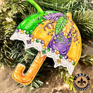 May include: A colorful Mardi Gras umbrella ornament with green, yellow, and purple sections. The ornament features a beaded border, decorative feathers, and a green ribbon for hanging. The handle is orange, and the design includes the text "DREZ Artwork & Design".