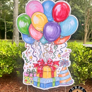 May include: A colorful birthday party yard sign featuring a bunch of balloons in various colors, including pink, red, yellow, green, blue, and purple. The balloons are tied together with ribbons and confetti. There are also three wrapped presents with bows and a party hat.