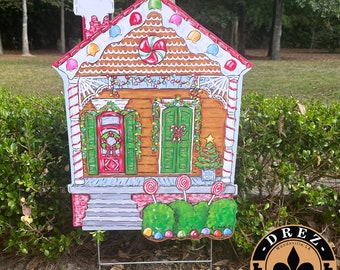 Könnte beinhalten: Eine Lebkuchenhaus-Garten Dekoration mit Zuckerstangen-Verzierungen, einer roten Tür mit einem Kranz und grünen Fensterläden. Das Haus hat ein weißes Dach mit Zuckerstangen-Lichtern und einer Zuckerstange auf der Veranda. Die Garten Dekoration wird von Drez Artwork & Design hergestellt, Made in the USA.