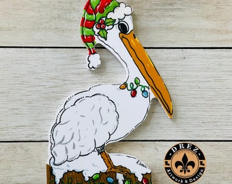 May include: A white pelican wearing a red and green Santa hat with holly and a string of lights around its neck. The pelican is standing on a brown log with more lights around it. The ornament is signed "T. Drez" and has a "Drez Artwork & Design" logo.