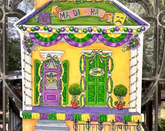 May include: A yellow and purple Mardi Gras house door hanger with green and gold beads, a purple door with a mask, and a green door with a wreath. The house has a porch with steps and a fence with green bushes.
