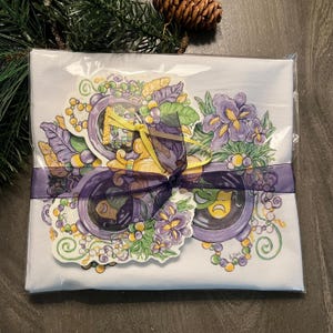 May include: A white gift set wrapped in clear plastic, tied with a purple ribbon and bow. The set features a colorful Mardi Gras design with purple irises, green leaves, and gold accents. Decorative stickers are included.