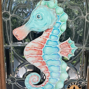 May include: A hand-painted wooden seahorse door hanger with a blue, green, and pink color scheme. The seahorse has a detailed design with scales and a curled tail. The hanger is decorated with a small fleur-de-lis and the text "Drez Artwork & Design".