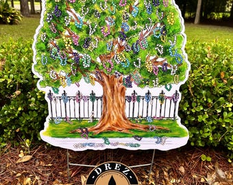 May include: A colorful yard sign featuring a tree with a brown trunk and green leaves adorned with beaded garlands. A white picket fence is at the base of the tree. The sign is set in a yard with green bushes and a logo at the bottom.